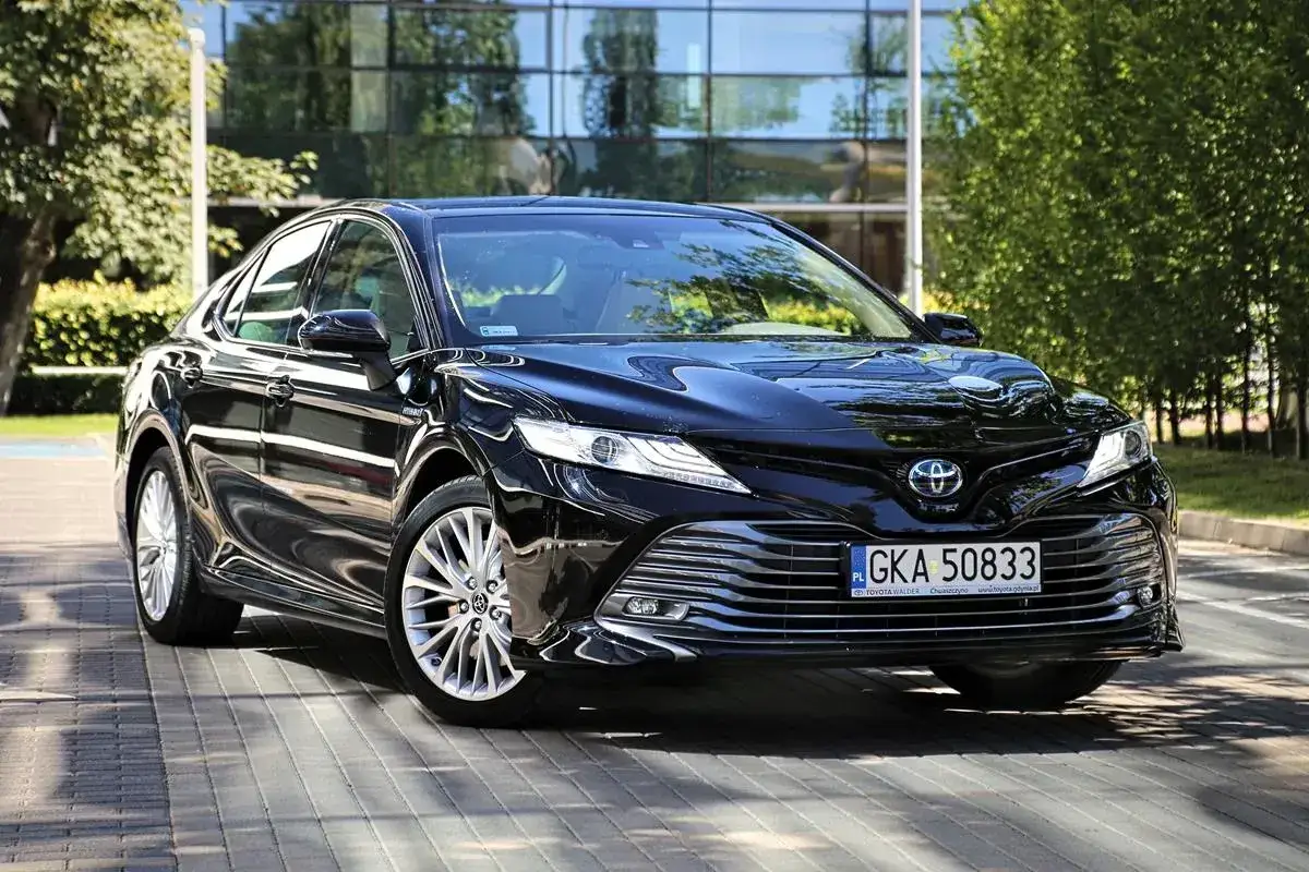 Elegancka, czarna Toyota Camry 2024 lśni na tle nowoczesnego budynku i zieleni.