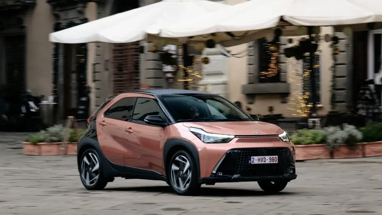 Różowy Toyota Aygo Cross z czarnym dachem przemierza urokliwe uliczki, błyszcząc w świetle.