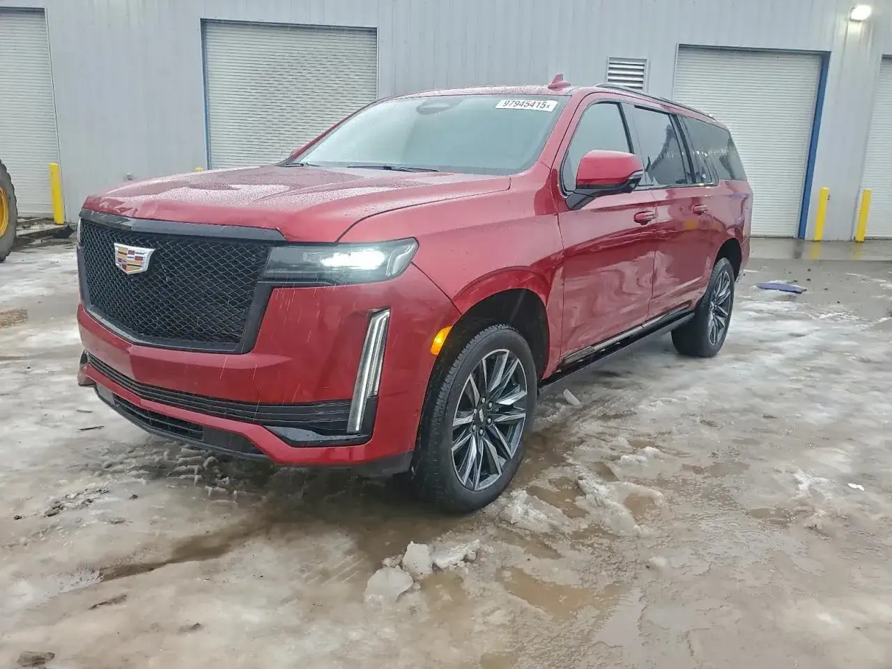 Czerwony Cadillac Escalade 2025 z czarną atrapą i felgami stoi na mokrym, zaśnieżonym placu przed budynkiem z bramami.