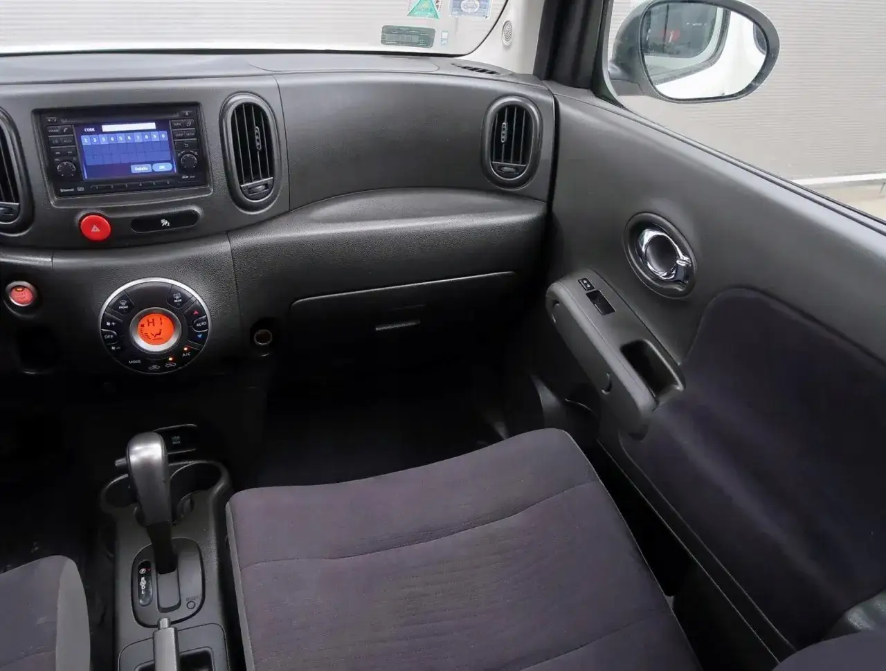 Wnętrze Nissan Cube: deska rozdzielcza z radiem, klimatyzacją i dźwignią zmiany biegów. Widok na drzwi pasażera.