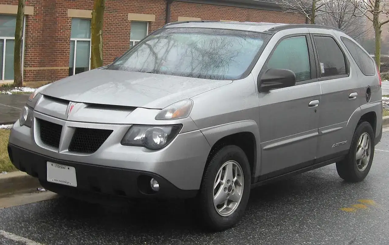 Srebrny Pontiac Aztek stoi na mokrej drodze przed ceglanym budynkiem.