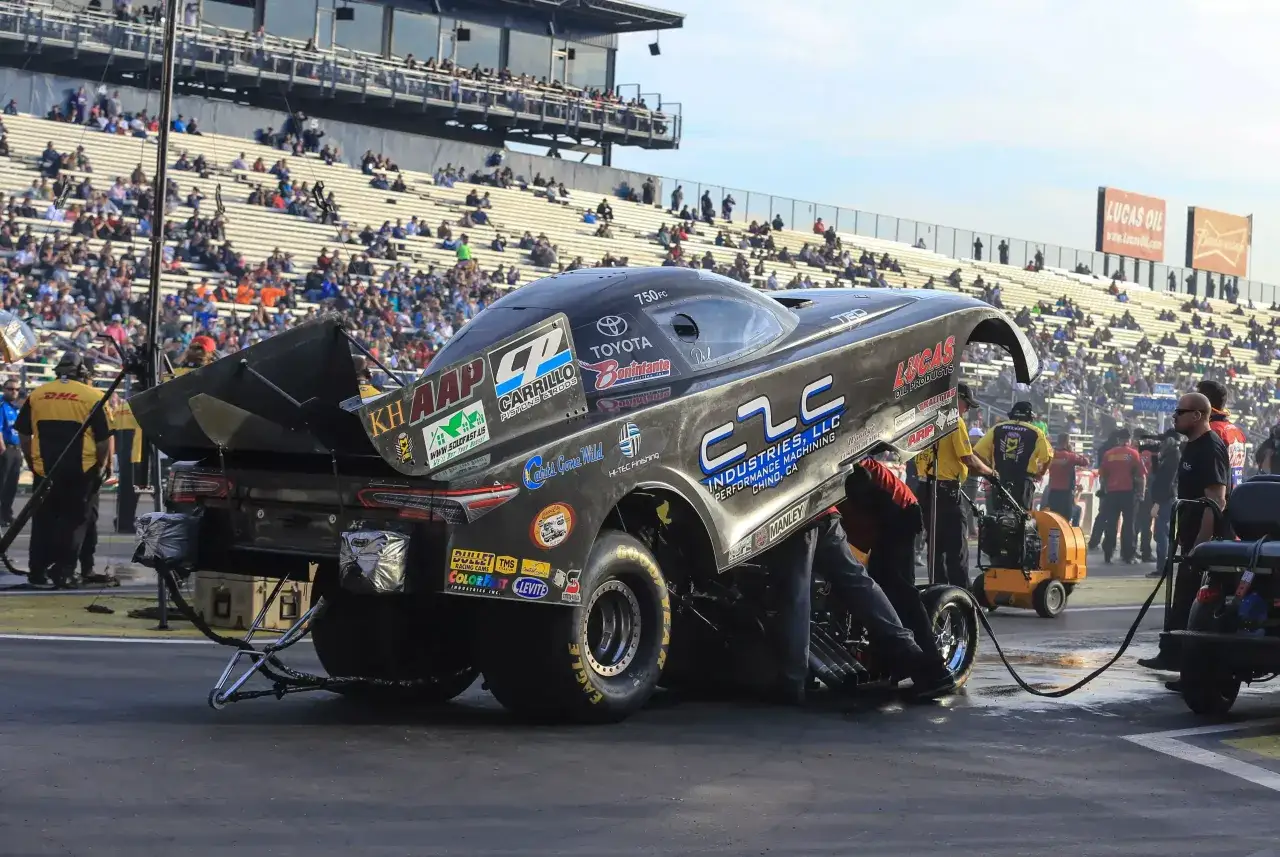 Czarny dragster z licznymi sponsorami, przygotowywany do wyścigu przez mechaników na tle trybun pełnych widzów.