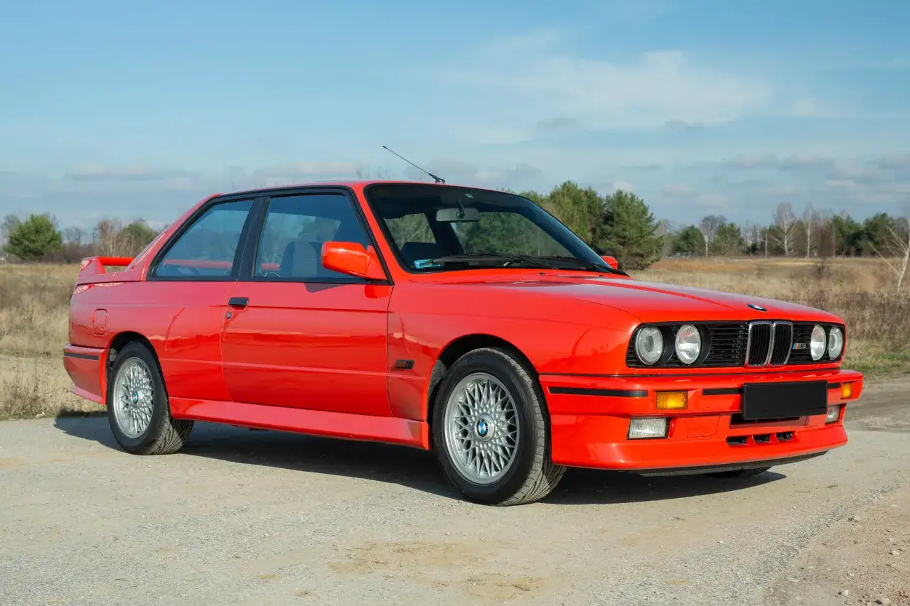 Klasyczne bmw e30 coupe w jaskrawym kolorze, zaparkowane na tle krajobrazu z drzewami.