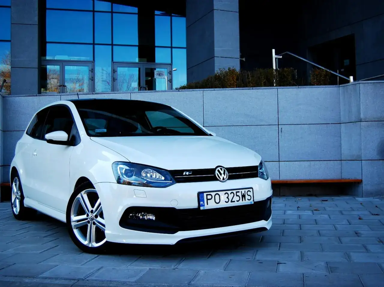Biały VW Polo 1.2 benzyna z czarnym dachem, zaparkowany przed nowoczesnym budynkiem.