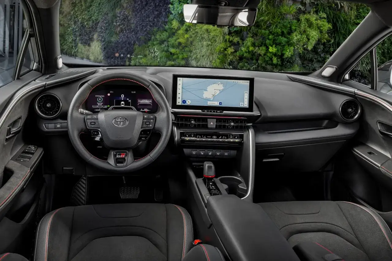 Wnętrze Toyoty C-HR 2023 z ekranem nawigacji i cyfrowymi zegarami. Czerwone przeszycia na kierownicy i fotelach dodają sportowego charakteru.