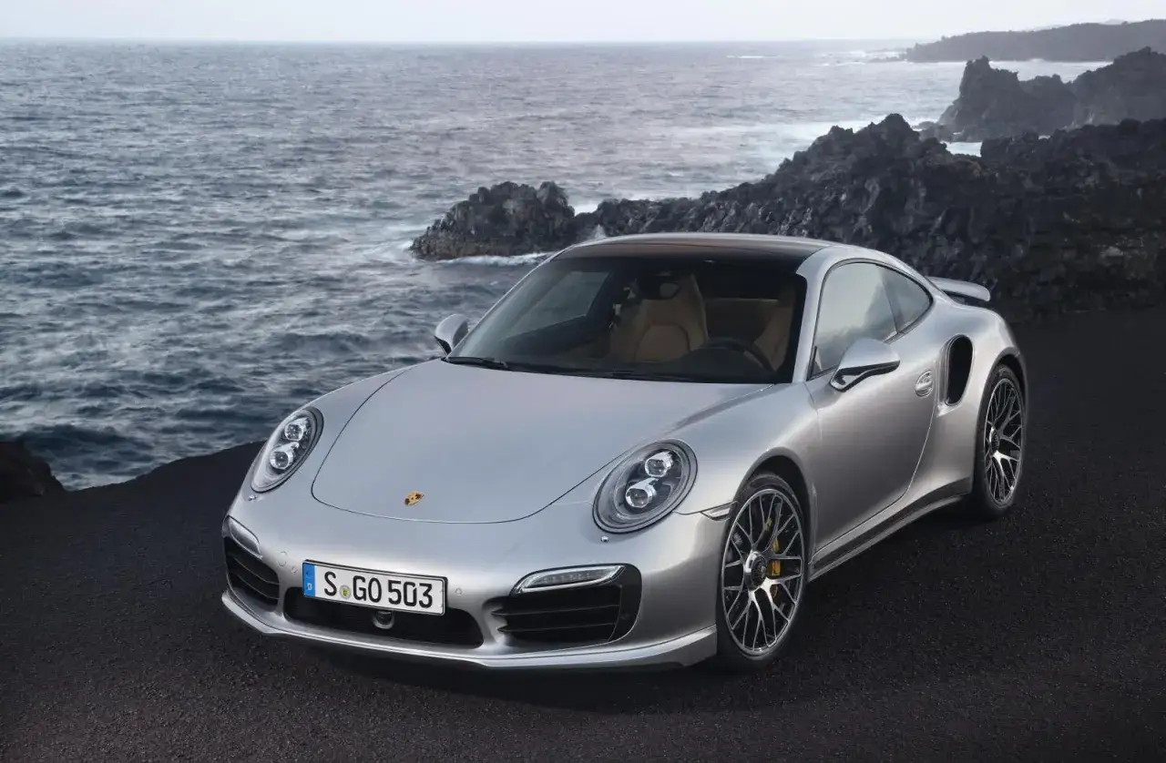 Srebrne Porsche 991 Turbo S z beżowym wnętrzem stoi na czarnej, wulkanicznej plaży. W tle widać wzburzone morze i skaliste wybrzeże.