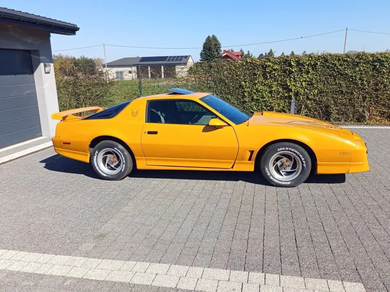 Żółty Pontiac Trans Am z 1987 roku, z charakterystycznym ptakiem na masce, zaparkowany przed domem.