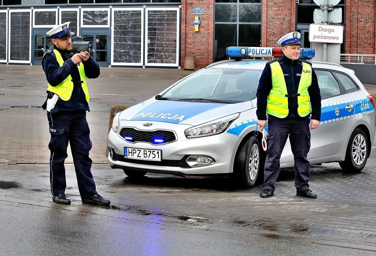 Policjanci z radarem mierzą prędkość. Jeden celuje, drugi stoi obok radiowozu. Kierowco, uważaj na przekroczenie prędkości o 20 km/h!