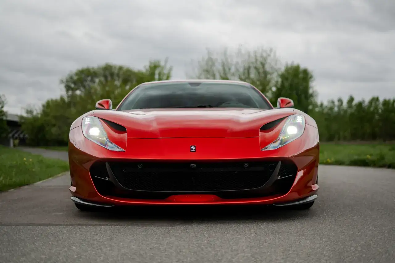 Czerwone Ferrari 812 Superfast z włączonymi światłami, zaparkowane na drodze pośród zieleni.