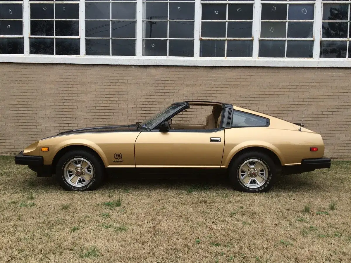 Złoty Datsun 280Z z czarnym dachem i pasem na masce, zaparkowany na trawie przed ceglanym budynkiem z dużymi oknami.