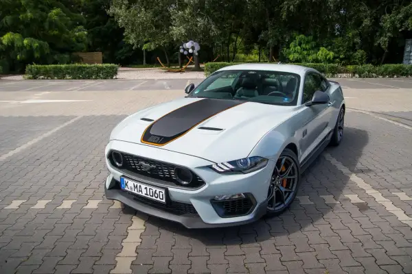 Biały Ford Mustang Mach 1 z czarnym pasem na masce i pomarańczowymi akcentami, zaparkowany na kostce brukowej.