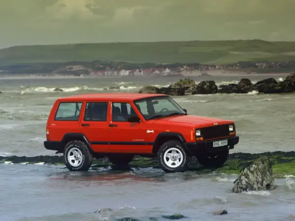 Czerwony Jeep ZJ na kamienistej plaży, z widokiem na morze i wzgórza w tle.
