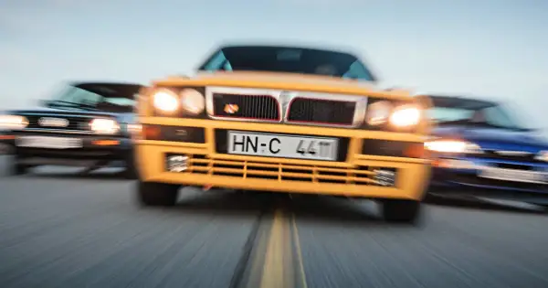 Żółta Lancia Delta Integrale w centrum, ścigana przez niebieski i czarny samochód.
