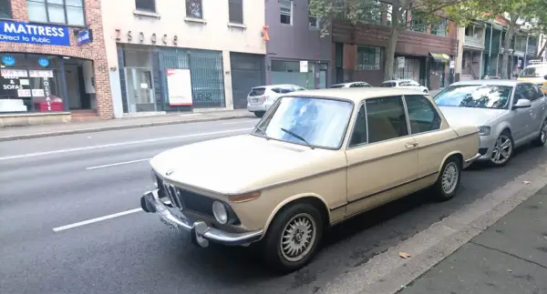 Klasyczny beżowy BMW E10 z lat 70. stoi zaparkowany przy ulicy, obok nowszego srebrnego samochodu.