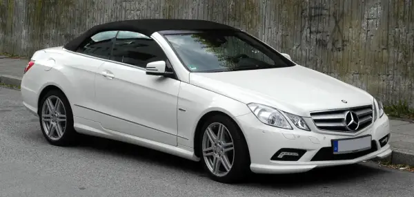 Biały kabriolet Mercedes W207 z czarnym dachem stoi na ulicy obok betonowej ściany.