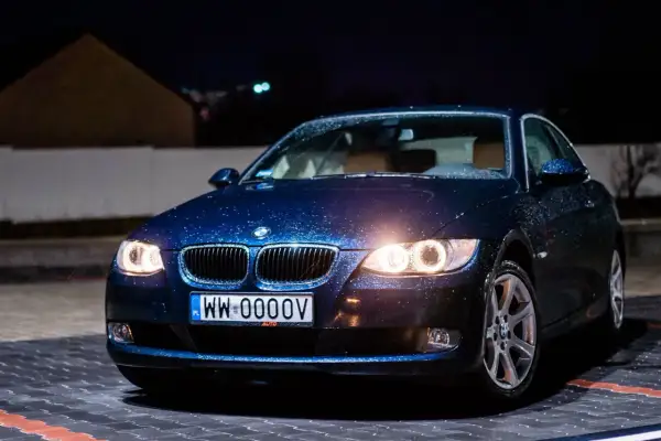Ciemnoniebieskie bmw 325i z kroplami deszczu na karoserii, zaparkowane nocą na mokrym parkingu.