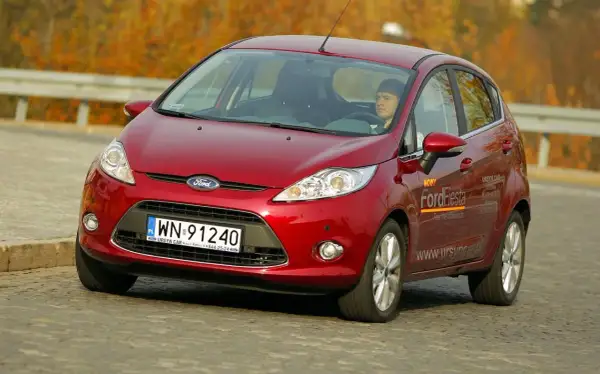 Czerwony Ford Fiesta 2009 z kierowcą za kierownicą, jadący po drodze.