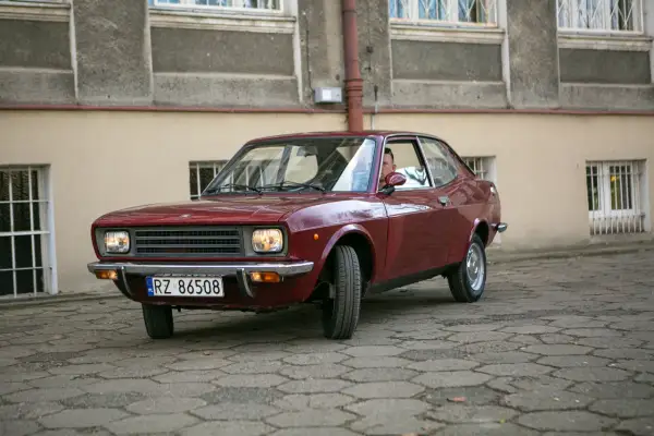 Czerwony fiat 128, klasyk z lat 70., zaparkowany na brukowanym dziedzińcu przed budynkiem.