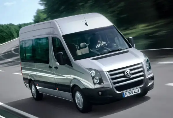 Srebrny Volkswagen Crafter jedzie po krętej drodze wśród drzew. Kierowca w białej koszuli prowadzi auto.
