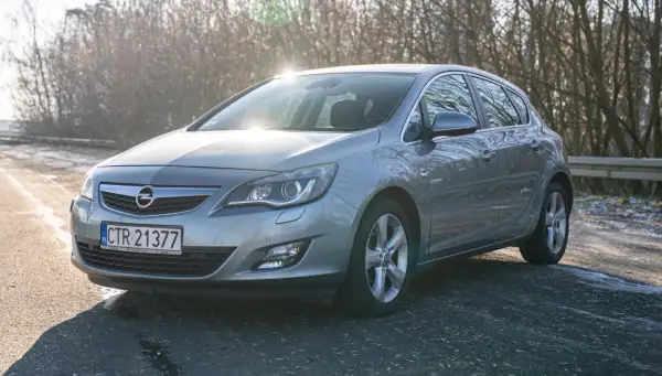 Szary Opel Astra J 1.6 benzyna na tle drzew.