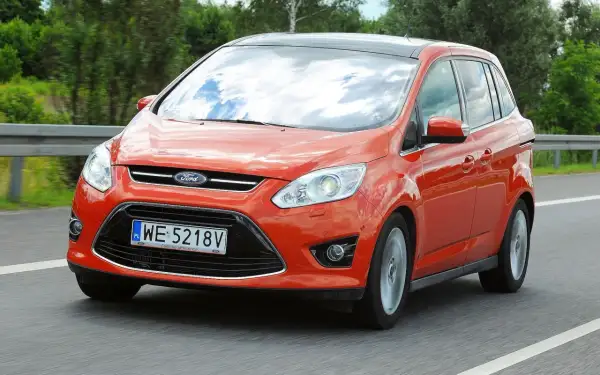 Pomarańczowy Ford C-Max 1.6 benzyna jedzie po drodze, odbijając chmury w szybie.
