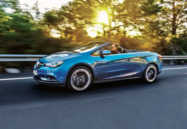 Niebieski Opel Cascada z otwartym dachem sunie po drodze, a słońce przebija się przez drzewa.