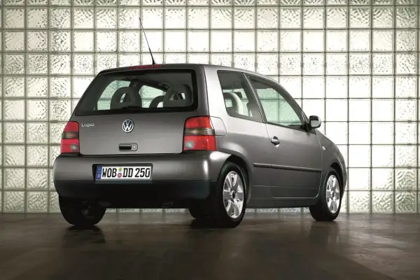 Szary Volkswagen Lupo stoi na ciemnej podłodze, na tle ściany z białych pustaków szklanych.